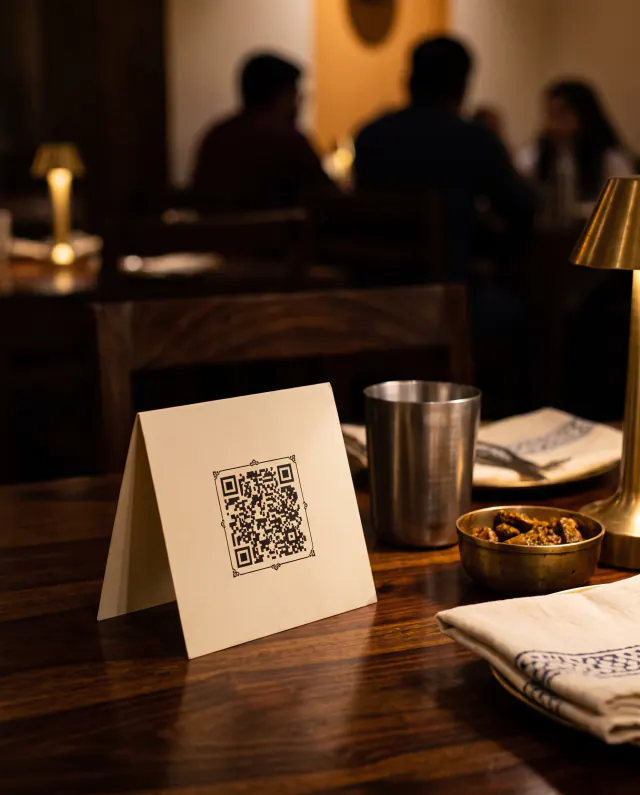 A small QR table tent on a wooden table beside a steel tumbler and a brass bowl of pickle.
